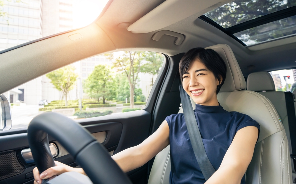 A woman with short black hair smiles broadly as she drives her new GMC SUV for the first time in her green neighborhood with the ceiling window open