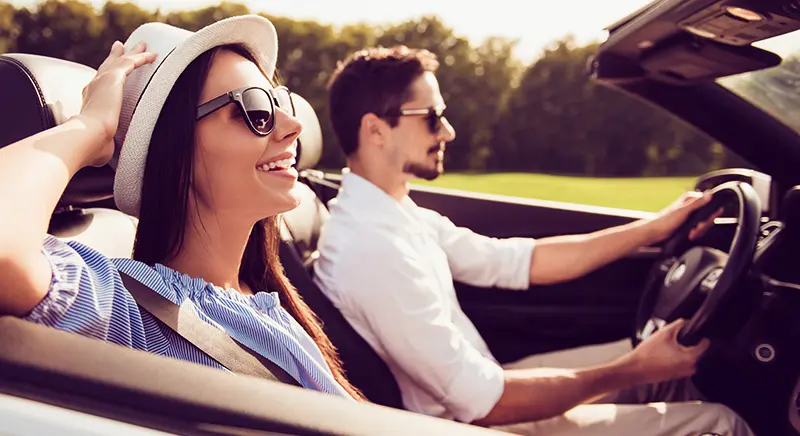 A couple smile & she holds her hat on as they enjoy a Summer Road Trips on a green country road